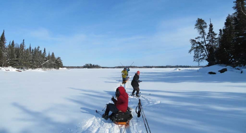 People sit and stand in a snowy landscape framed by evergreen trees